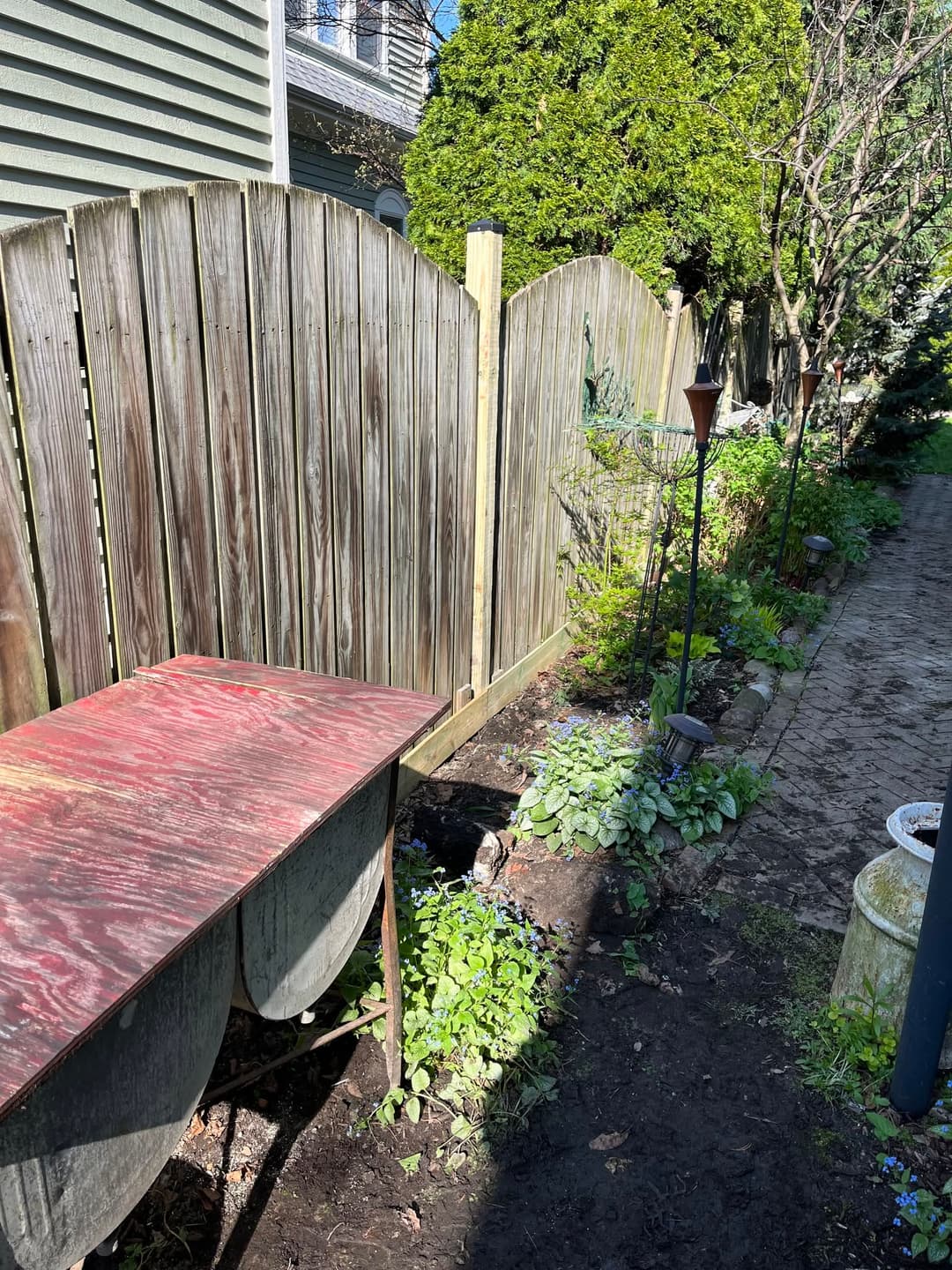 Wooden fence along a garden path with plants, lanterns, and a weathered table.