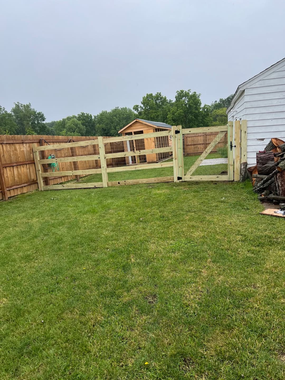 Wooden fence gate opening to a grassy yard with a shed and trees in the background.