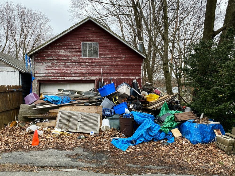 Cluttered garage with debris, tarps, and discarded items, surrounded by trees and bare ground.