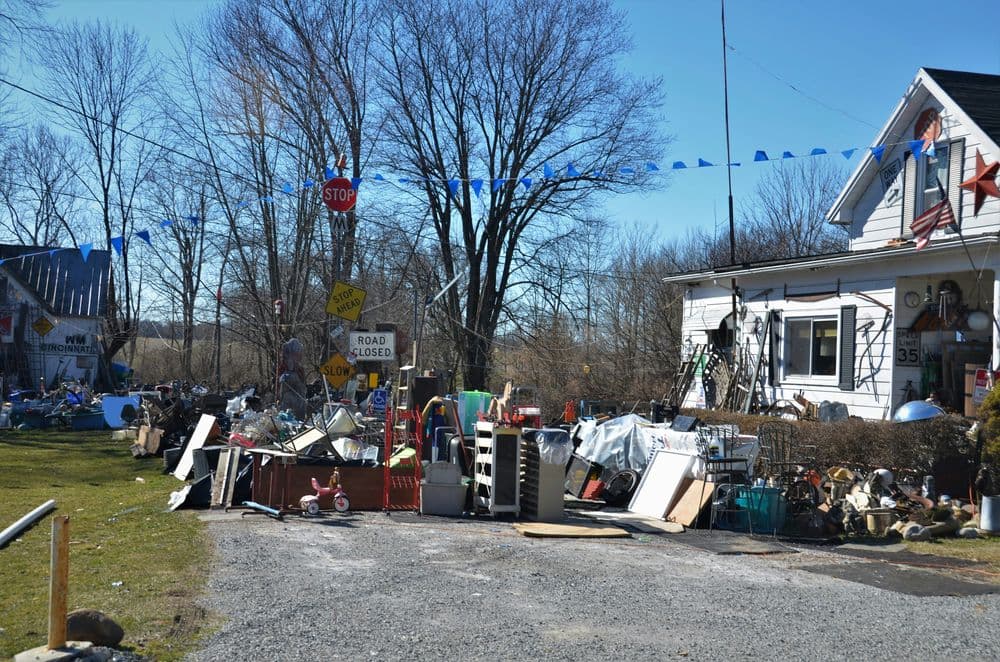 Cluttered yard filled with discarded items, road signs, and a white house in background.