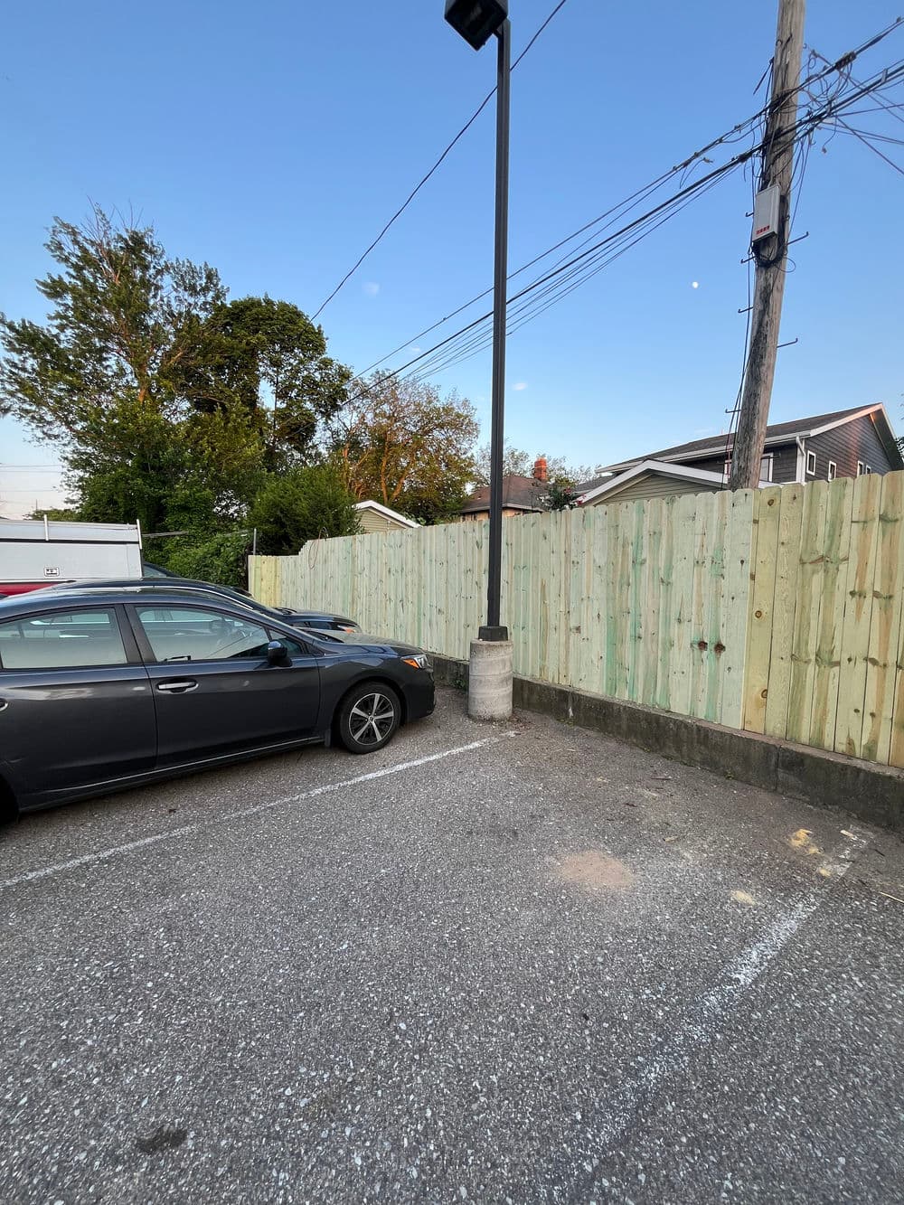 Black car parked next to a green wooden fence in an outdoor lot during sunset.