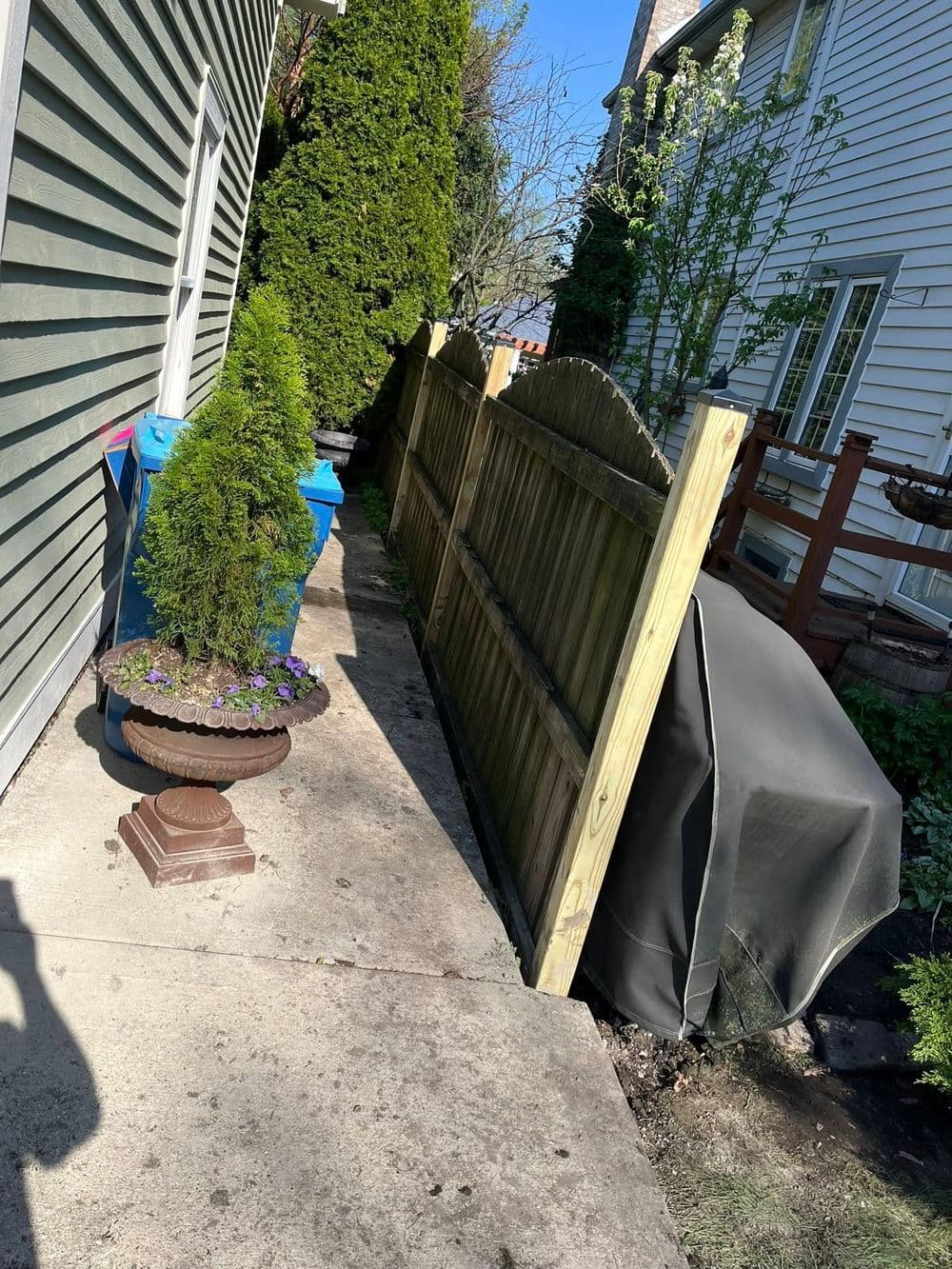 Narrow backyard pathway with a wooden fence, potted plants, and a grill cover.